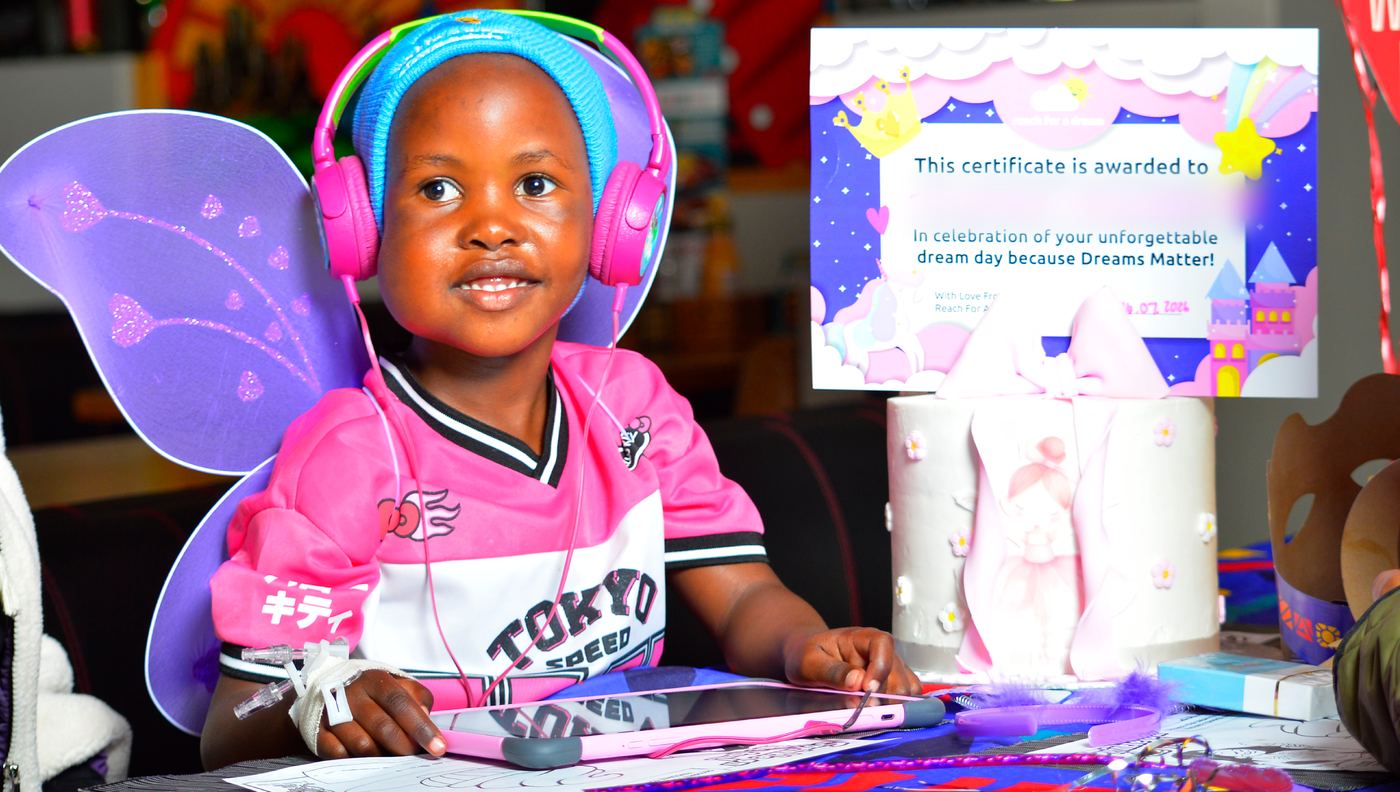 A young girl in fairy wings and headphones at her dream-day table, a decorated cake beside her