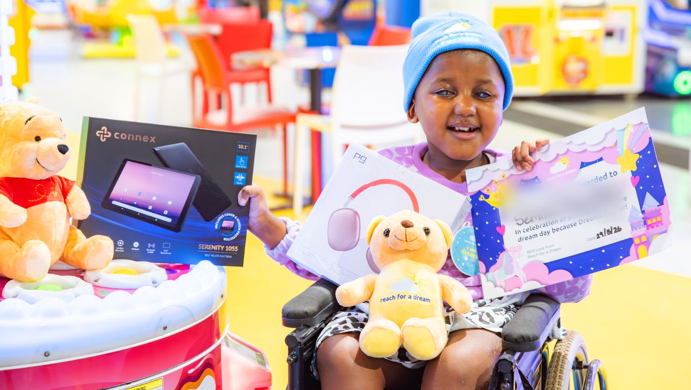 A girl in a wheelchair holding up a tablet, headphones and a teddy bear